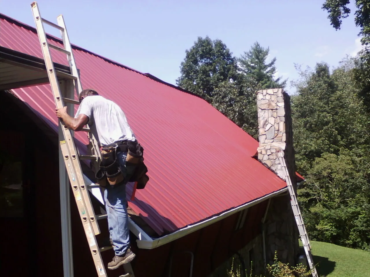 Roofer on a ladder beside a red metal roof and stone chimney — Soffit & Fascia Repair by Roofing Brothers in West Wardsboro